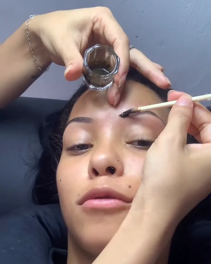 Mãos da profissional aplicando produto na sobrancelha de uma cliente deitada, de olhos semicerrados.