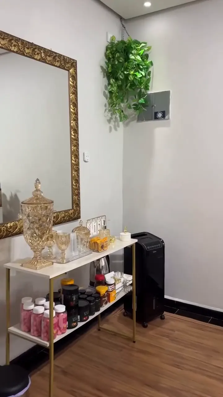 Interior do studio: espelho de moldura dourada, planta pendente e estante com potes dourados sobre piso de madeira.